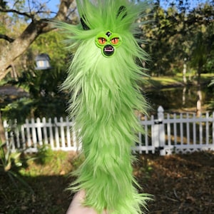 May include: A long, green plush toy with a fuzzy texture. The toy has a face with orange eyes and a black mouth. It has black ears and a spiky head. The toy is held in front of a blurred outdoor background.