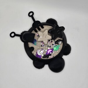 May include: Black silicone sensory toy shaped like a turtle with a sand-like texture inside. The toy includes a small shovel, rake, and star-shaped mold. The sand is decorated with glitter and small plastic stars, moons, and planets.