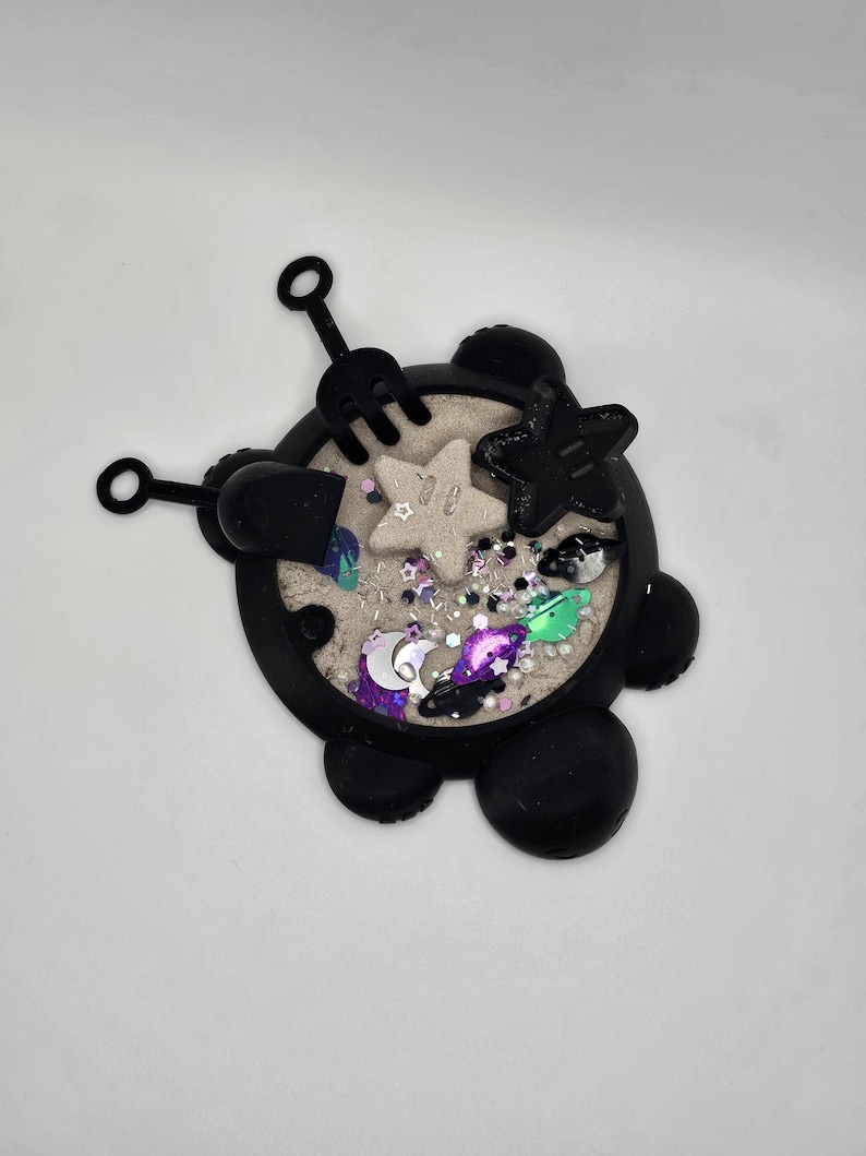 May include: Black silicone sensory toy shaped like a turtle with a sand-like texture inside. The toy includes a small shovel, rake, and star-shaped mold. The sand is decorated with glitter and small plastic stars, moons, and planets.