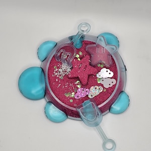 May include: A blue plastic turtle-shaped sandbox with pink sand and glitter. It includes a small shovel and two star-shaped molds.