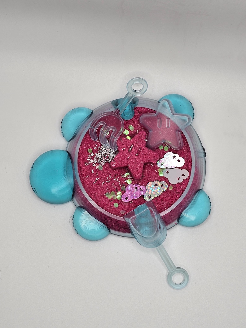 May include: A blue plastic turtle-shaped sandbox with pink sand and glitter. It includes a small shovel and two star-shaped molds.