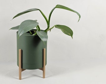 May include: A green potted plant with large, light green leaves in a dark green planter with a gold metal stand.