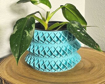 May include: A light blue geometric planter with a white and green variegated plant. The planter is made of a textured material and has a unique design. The plant is in a pot and is sitting on a wooden surface.