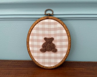 May include: An oval-shaped embroidery hoop with a brown wooden frame. The fabric inside features a light brown and white gingham pattern, with a brown teddy bear stitched in the center. The hoop has a metal clasp at the top.