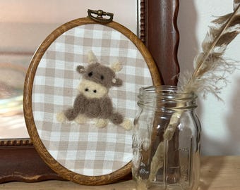 May include: A brown and white gingham fabric with a brown felt cow embroidered on it. The fabric is in a wooden embroidery hoop.