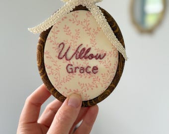 May include: A round, embroidered hoop with the name "Willow Grace" in burgundy thread. The fabric has a pink floral pattern. The wooden hoop is topped with a cream-colored lace bow. A decorative item.