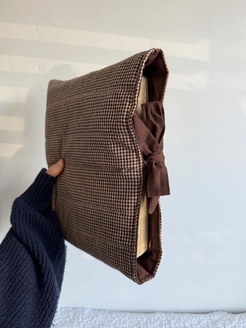 Op de afbeelding: Een bruin en tan geruite boekomslag met een donkerbruine striksluiting. De omslag is gewatteerd en heeft een zachte, gewatteerde textuur. De boekomslag is ontworpen om het uiterlijk van een boek te beschermen en te verbeteren.