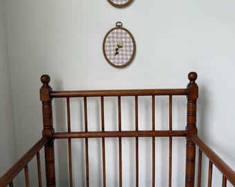 May include: A wooden crib with a floral patterned quilt and two framed embroidery hoops hanging on the wall. The embroidery hoops have a gingham pattern and a floral design.