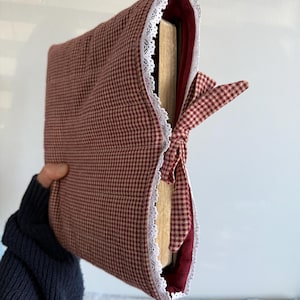 May include: A burgundy and white gingham book cover with a white lace trim. The cover is tied with a matching gingham bow. The interior is a solid burgundy color. The book cover is designed to protect a book.
