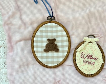 May include: Two embroidered wall hangings. One features a brown teddy bear on a tan and white checkered background, framed in wood, with a blue ribbon. The other has the name "Willow Grace" on a floral background.