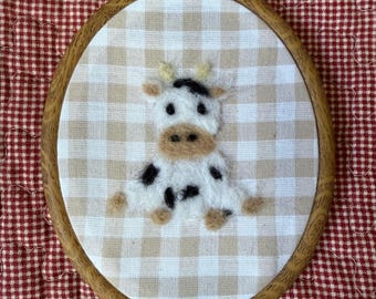 May include: A handcrafted embroidery hoop featuring a charming cow design. The cow has black spots, a beige muzzle, and a white body. The background is a beige and white checkered pattern, set within a wooden hoop.