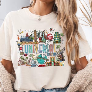 May include: Ivory-colored t-shirt featuring a colorful graphic of Universal Studios attractions. The design includes the words "UNIVERSAL STUDIOS" and various illustrations of theme park elements. The shirt is a comfortable, casual style.