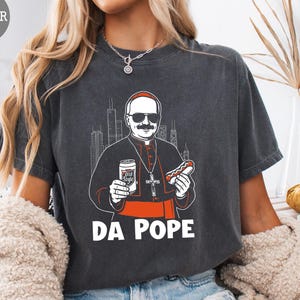 May include: Dark gray t-shirt featuring a cartoon illustration of a pope-like figure with sunglasses, holding a beer and a hot dog. The text "DA POPE" is printed below the image. The shirt has a vintage, worn look.