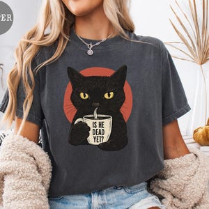 May include: Dark gray t-shirt featuring a black cat with yellow eyes holding a mug. The mug has the text "IS HE DEAD YET?" printed on it. The cat is set against a red circle. A casual, Halloween-themed tee.