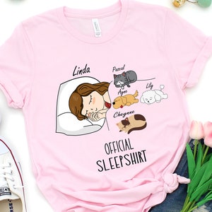 May include: Pink t-shirt with a cartoon illustration of a woman sleeping with four pets. The text "Linda" is above the woman's head. The text "Pascal, Ayne, Lily, Cheyenne" is above each pet. The text "Official Sleepshirt" is below the illustration.