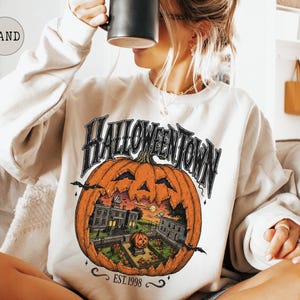 May include: A sand-colored crewneck sweatshirt featuring a large graphic print of a carved pumpkin containing a spooky town scene. The text HALLOWEENTOWN is printed in a gothic font above the pumpkin, with EST. 1998 printed in smaller text below.