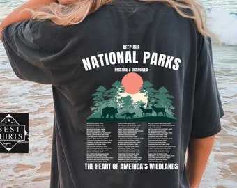 May include: A dark gray t-shirt with a graphic print of a forest scene and the text "Keep our National Parks Pristine & Unspoiled." The text "The Heart of America's Wildlands" is printed below the graphic. A list of national parks is printed below the text.