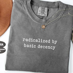 May include: A gray Comfort Colors t-shirt with the text "radicalized by basic decency" in white. A gold necklace with a pendant is draped on the shirt. A brown hat and decorative leaves are in the background.