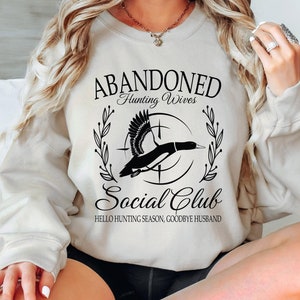 May include: Cream-colored sweatshirt with the text "ABANDONED Hunting Wives Social Club." The design features a flying duck silhouette within a circle, with the words "HELLO HUNTING SEASON, GOODBYE HUSBAND" below. The sweatshirt has long sleeves.