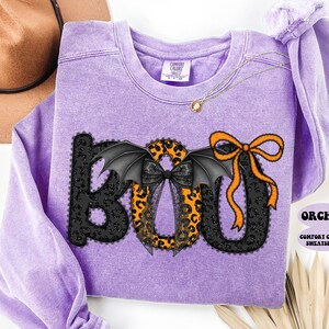 May include: A lavender Comfort Colors sweatshirt with a Halloween-themed "BOO" design. The "B" and "O" are black with lace trim, and the "O" has bat wings and a leopard print. The "O" has an orange bow.
