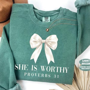 May include: Light green sweatshirt with a large white bow graphic and the text "SHE IS WORTHY PROVERBS 31". The sweatshirt has a crew neck and long sleeves. A gold necklace is draped on the front.