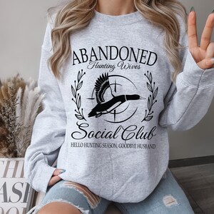 May include: A light gray sweatshirt with the text "ABANDONED Hunting Wives Social Club" in a black script font. A black and white graphic of a duck is in the center. The text "HELLO HUNTING SEASON, GOODBYE HUSBAND" is below the graphic.