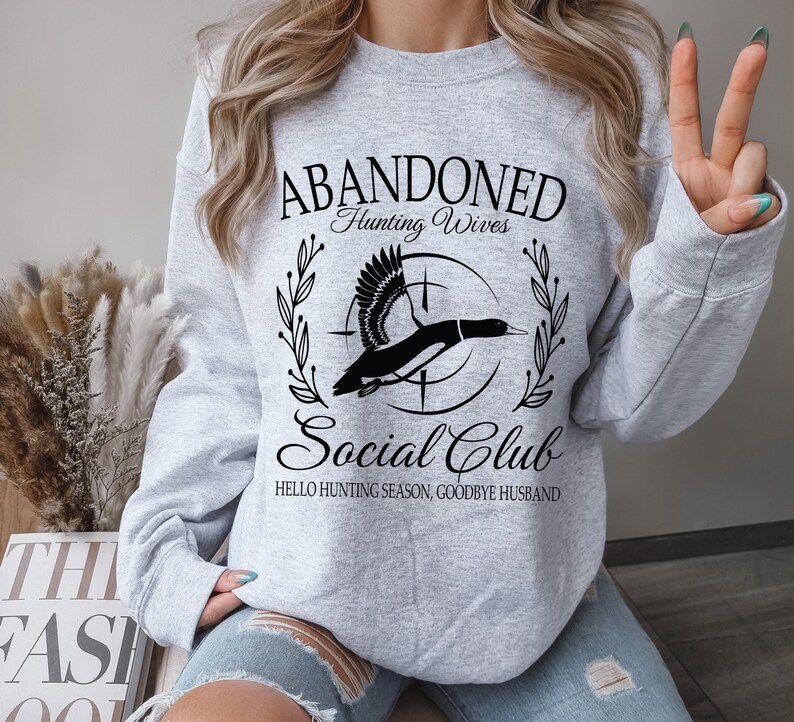 May include: A light gray sweatshirt with the text "ABANDONED Hunting Wives Social Club" in a black script font. A black and white graphic of a duck is in the center. The text "HELLO HUNTING SEASON, GOODBYE HUSBAND" is below the graphic.