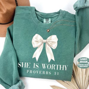 May include: Light green sweatshirt with a large white bow graphic and the text "SHE IS WORTHY PROVERBS 31". The sweatshirt has a crew neck and long sleeves. A gold necklace is draped on the front.