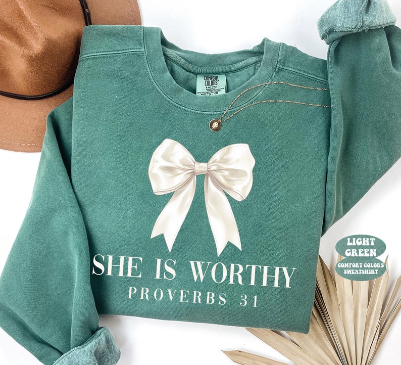 May include: Light green sweatshirt with a large white bow graphic and the text "SHE IS WORTHY PROVERBS 31". The sweatshirt has a crew neck and long sleeves. A gold necklace is draped on the front.