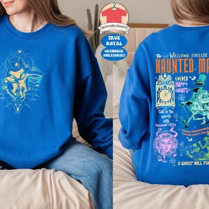 May include: A blue sweatshirt with a graphic print of the Haunted Mansion attraction from Disneyland. The print features the text "Welcome Foolish Mortals Haunted Mansion" and various characters from the attraction, including the Ghost Host.