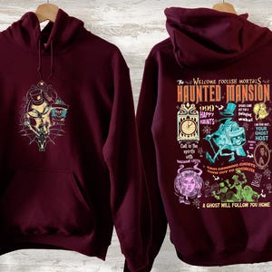 May include: Burgundy hooded sweatshirt with a graphic print of the Haunted Mansion attraction at Disneyland. The print features the text "The Welcome Foolish Mortals Haunted Mansion" and various characters from the attraction.