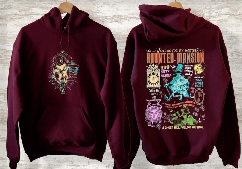 May include: Burgundy hooded sweatshirt with a graphic print of the Haunted Mansion attraction at Disneyland. The print features the text "The Welcome Foolish Mortals Haunted Mansion" and various characters from the attraction.