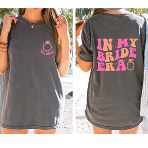 May include: A gray t-shirt with a pink and orange graphic that says "In My Bride Era" with a diamond ring. The front of the shirt has a small diamond ring graphic with the text "Mrs. Johnson".