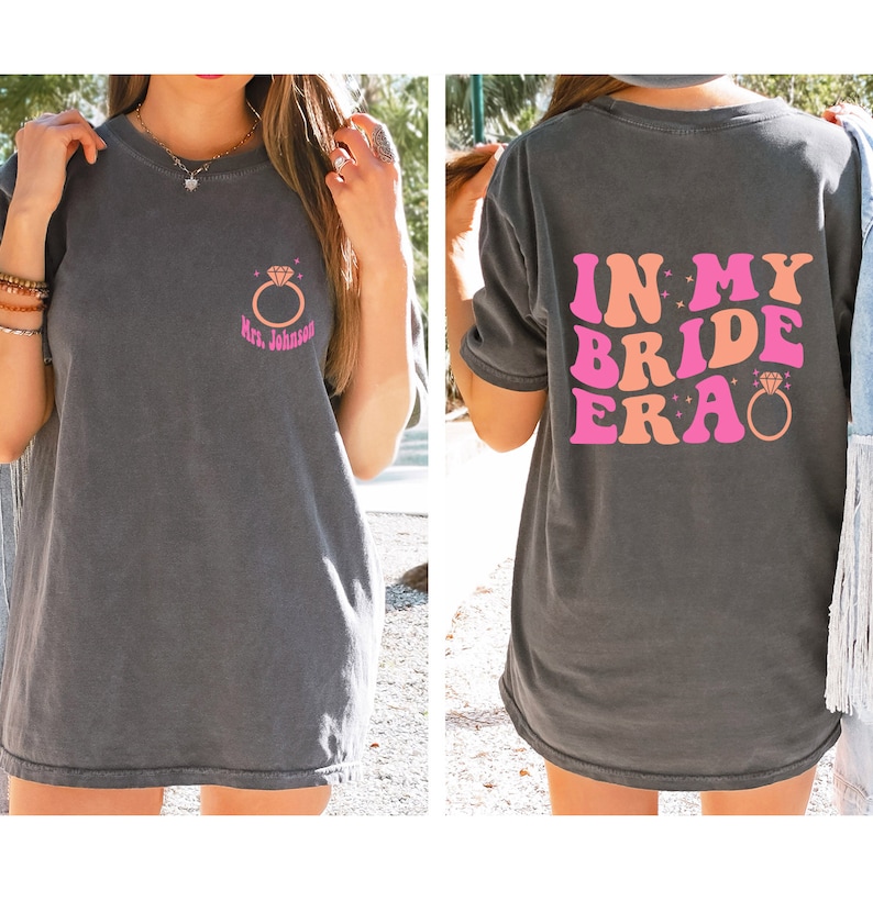 May include: A gray t-shirt with a pink and orange graphic that says "In My Bride Era" with a diamond ring. The front of the shirt has a small diamond ring graphic with the text "Mrs. Johnson".