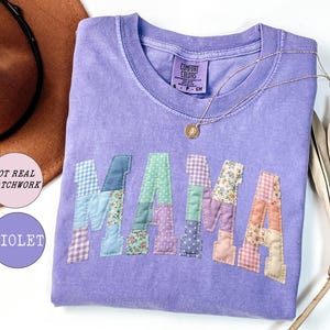 May include: A violet Comfort Colors t-shirt with the word "MAMA" appliqued across the front in a patchwork design. The letters are made of various floral and patterned fabrics. A gold necklace with a pendant rests on the shirt.