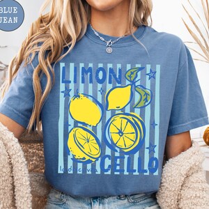 May include: Blue jean t-shirt with a graphic of yellow lemons and the words "LIMONCELLO" in blue. The design includes blue vertical stripes and star accents. The shirt is a casual, comfortable style.