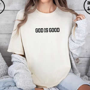 Puede incluir: Una camiseta blanca con el texto "GOD IS GOOD" impreso en negro.