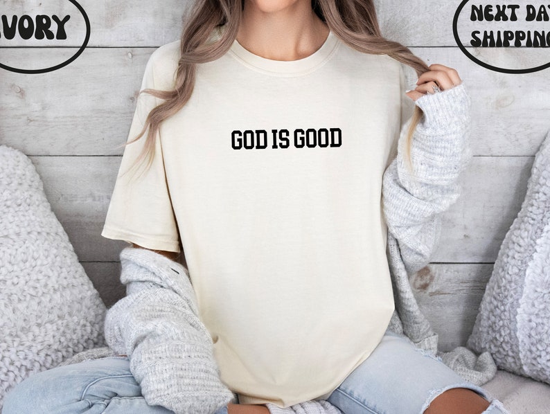 Puede incluir: Una camiseta blanca con el texto "GOD IS GOOD" impreso en negro.