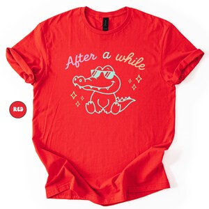 Puede incluir: Camiseta roja con el texto "After a while" sobre un cocodrilo de dibujos animados con gafas de sol. El cocodrilo est&aacute; delineado en azul claro, con texto y estrellas a juego. Un c&iacute;rculo rojo con la palabra "RED" est&aacute; en la esquina.