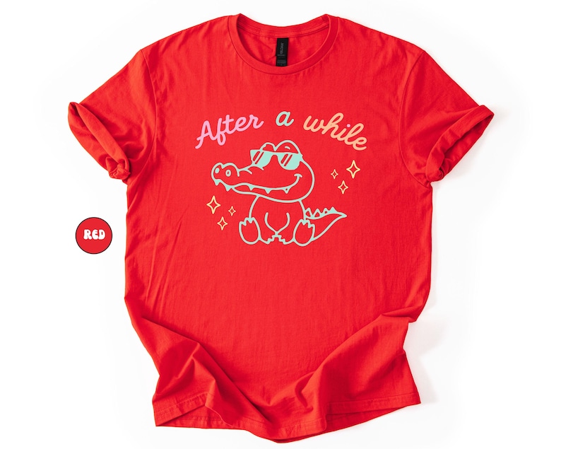 Puede incluir: Camiseta roja con el texto "After a while" sobre un cocodrilo de dibujos animados con gafas de sol. El cocodrilo est&aacute; delineado en azul claro, con texto y estrellas a juego. Un c&iacute;rculo rojo con la palabra "RED" est&aacute; en la esquina.