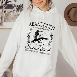 May include: White hoodie with a front graphic that reads "ABANDONED Hunting Wives Social Club." The graphic features a flying duck inside a circle with text that says "Hello Hunting Season, Goodbye Husband."