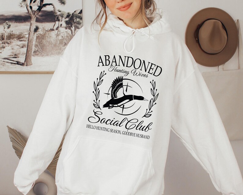 May include: White hoodie with a front graphic that reads "ABANDONED Hunting Wives Social Club." The graphic features a flying duck inside a circle with text that says "Hello Hunting Season, Goodbye Husband."
