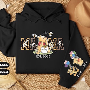 May include: Black hooded sweatshirt with a Winnie the Pooh design. The text "Mama Bear Est. 2025" is printed on the front. The sleeves feature Winnie the Pooh characters with balloons.