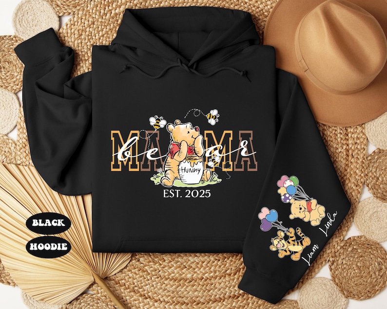 May include: Black hooded sweatshirt with a Winnie the Pooh design. The text "Mama Bear Est. 2025" is printed on the front. The sleeves feature Winnie the Pooh characters with balloons.