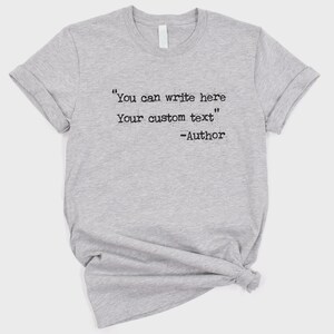 Może przedstawiać: Szara koszulka z podwiniętymi rękawami. Koszulka zawiera tekst "You can write here Your custom text" a następnie "-Author" czarną czcionką w stylu maszyny do pisania. Koszulka jest zawiązana na dole.