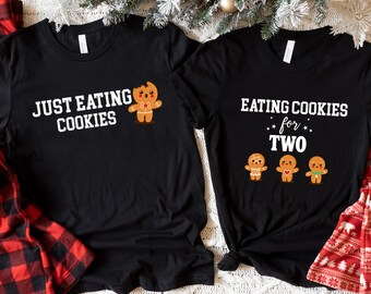May include: Two black t-shirts with a Christmas theme. One shirt says "Just Eating Cookies" with a gingerbread man graphic. The other shirt says "Eating Cookies for Two" with three gingerbread people graphics.