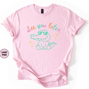 Puede incluir: Camiseta rosa claro con un cocodrilo de dibujos animados con gafas de sol. El texto "See you later" est&aacute; escrito encima del cocodrilo en colores pastel. La camiseta tiene mangas enrolladas y cuello redondo.