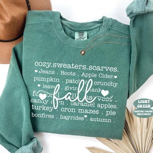 May include: A light green Comfort Colors sweatshirt with the word "fall" in a cursive font. The shirt features a list of fall-themed words in white, including "cozy," "sweaters," and "pumpkin." A gold necklace is draped across the shirt.