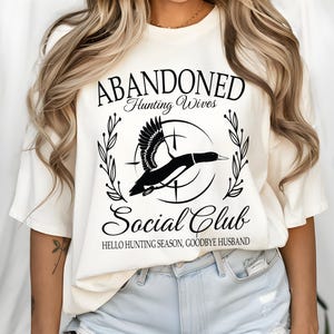 May include: Cream-colored t-shirt with a black graphic design. The design features a flying duck within a circle, surrounded by text that reads "ABANDONED Hunting Wives Social Club" and "HELLO HUNTING SEASON, GOODBYE HUSBAND."