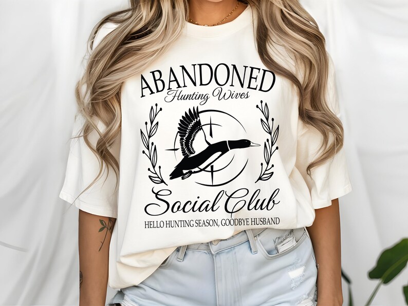 May include: Cream-colored t-shirt with a black graphic design. The design features a flying duck within a circle, surrounded by text that reads "ABANDONED Hunting Wives Social Club" and "HELLO HUNTING SEASON, GOODBYE HUSBAND."